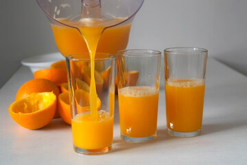 Woman is pouring fresh orange juice from electric juicer into three glasses, closeup view. Healthy vegan ripe fruit juice, natural vitamins from food. Vegan healthy organic nutrition.