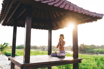 Side view of calm female in sportive tracksuit sitting in lotus pose and keep praying during morning meditation near rice fields, young Caucasian woman relaxing during yoga concentration in Thailand