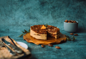 Homemade cake with chocolate and cream on round wooden board next to a plate with a fork and knife and a mug filled with nuts on dark background. Home baking