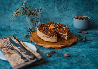 Homemade cake with chocolate and cream on round wooden board next to a plate with a fork and knife and a mug filled with nuts on dark background. Home baking