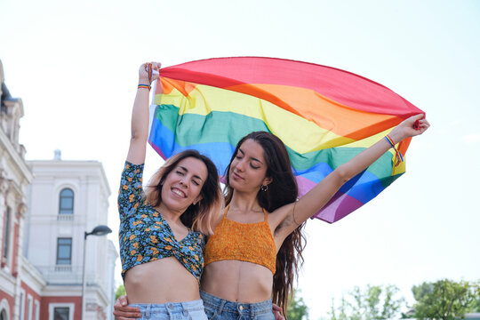 Young lesbian couple smiling and holding a rainbow flag.