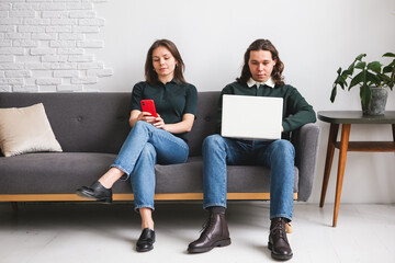 Couple sitting on the sofa with notebook and phone. Man and woman communicating on the electronic gadgets. Social isolation, distant learning students