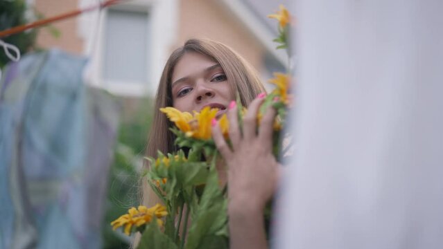 Slim charming woman posing with bouquet of sunflowers outdoors as clean laundry hanging at front. Portrait of beautiful Caucasian lady looking at camera bragging flowers and fresh laundered clothes