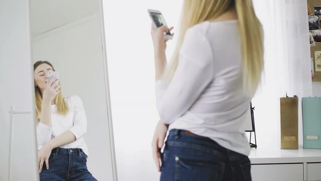 A young woman looks in the mirror, preens herself, takes pictures of herself on her phone in the mirror, adjusts her clothes and hairstyle. He smiles and likes himself.