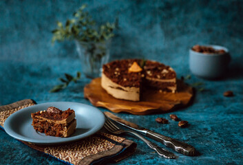Homemade cake with chocolate and cream on round wooden board, plate with piece of cake next to fork and knife and mug filled with nuts on dark background. Home baking
