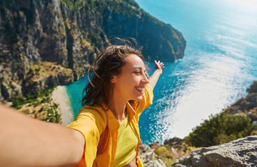 Positive tourist woman taking picture outdoors for memories, making selfie on top of cliff with valley mountains view, sharing travel adventure journey - 484225621