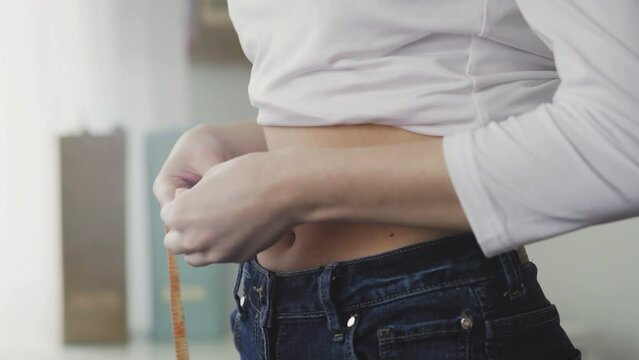 A young woman stands in front of a mirror and measures the circumference of her waist with a tape measure. Close-up
