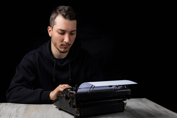 Young handsome man in black hoodie is typing on a typewriter, on black background in low key