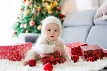 Little baby boy in white knitted onesie, playing with and opening presents at home
