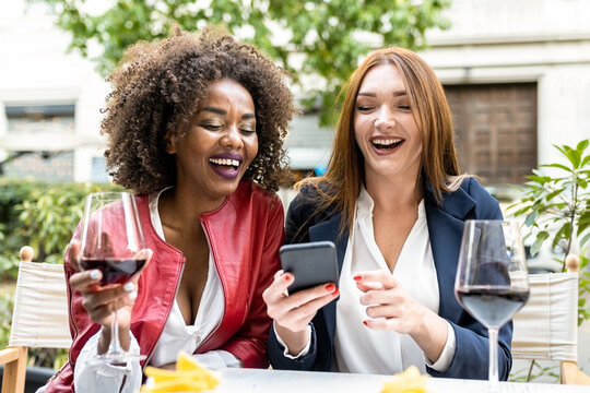 two adult woman having fun during happy hour after work, colleagues laughing while looking at smart phone, social network and mobile app for tech addicted people - Powered by Adobe