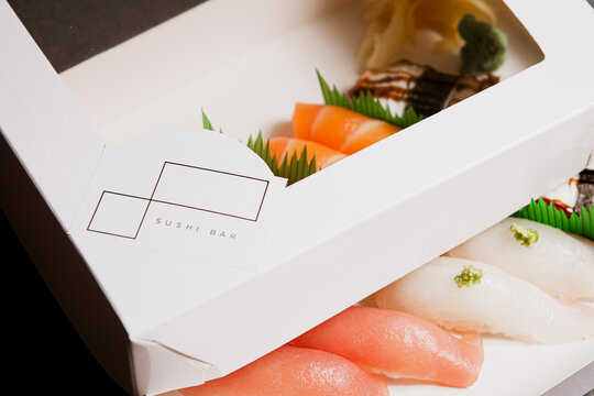 Sushi to go concept. Top view of takeaway box with various sushi rolls in a withe ecologic paper box. Hand holding box. - Powered by Adobe