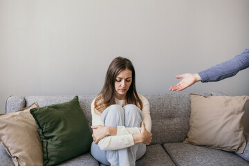 a sad woman is sitting on the couch and hugging herself. An outstretched helping hand. Domestic violence