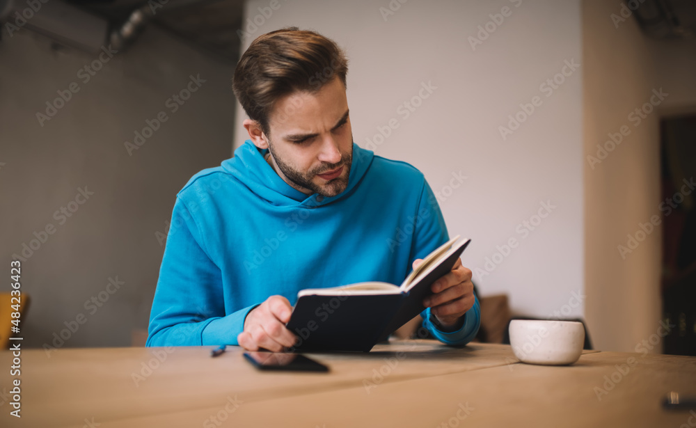 Poster caucasian male student reading informative notes from education textbook while learning in coffee sh