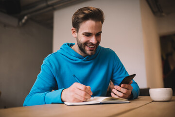 Happy Caucasian male student enjoying leisure time for creating content ideas using cellular gadget, cheerful hipster guy writing in education textbook while checking browsed mobile information