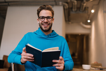 Portrait of cheerful student in optical eyewear holding education textbook smiling at camera while...