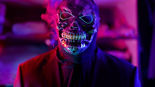 a man in a terrible glowing mask of death lets out clouds of smoke from under it in a neon flashing light. close-up. the dark key