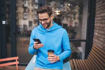 Cheerful hipster guy with takeaway coffee to go enjoying mobile messaging during coffee time on weekend, happy Caucaisan man in classic eyeglasses connecting to 4g wireless for making online booking