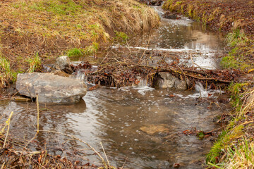 A rock in a stream in front of a small reservoir. A stream passes through a meadow of green grass. Rocks have created a natural dam. A large block of rock is in front of this dam.
