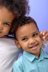 Two happy black children posing at camera and cheerfully smiling over purple studio background, cute american brother and sister having fun together, wearing casual shirts. childhood concept