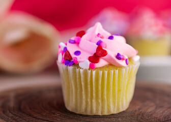 A vanilla valentine's day cupcake on a wooden slab and tulips and additional cupcakes blurred in the background.  Pink background.