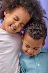 Two happy black sister and brother having fun while standing isolated over purple background, portrait of black children in casual clothes laughing, smiling together. human emotions concept