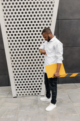 Stylish dressed businessman standing near the office, looking at wristwatch, waiting for the client. African american male employee holding a laptop, checking time, concept of business and timekeeping