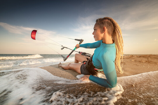 Attractive caucasian woman kitesurfer in a neoprene wetsuit and with dreadlocks on her head is resting lying on a sandy beach on the shore holding her kite in the wind