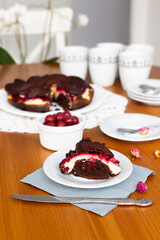 Piece of homemade brownie pie with cream cheese and cherry filling on white plate and grey napkin on wooden table