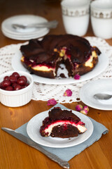 Piece of homemade brownie pie with cream cheese and cherry filling on white plate and grey napkin on wooden table