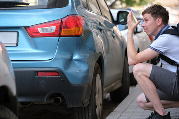 Sad male driver sitting on street side shocked looking at his dented car parked at street side. Road safety and vehicle insurance concept