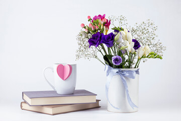 Cup with  paper heart, beautiful flowers  in vintage ceramic vase with ribbon, and books on white background. Romantic morning concept background with copy space.