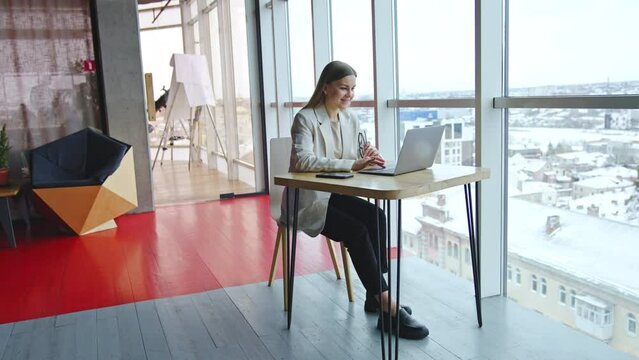 Young woman at her workplace in light spacious office. Business lady sitting at desk and working at laptop. Cityscape at the backdrop.
