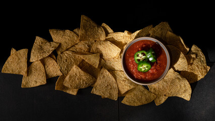 Yellow corn tortilla chips with red salsa topped with green sliced Jalapenos on dark background. - obrazy, fototapety, plakaty