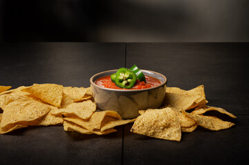 Yellow corn tortilla chips with red salsa topped with green sliced Jalapenos on dark background.