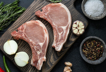 Raw steaks on a cutting board with salt, rosemary, onion, hot pepper and garlic, top view. Fresh pork steaks with spices and herbs on a dark background.