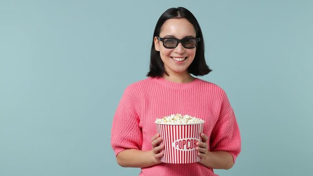Fun young woman of Asian ethnicity 20s wears pink shirt 3d glasses hold bucket of popcorn look aside on workspace area copy space mock up isolated on plain pastel light blue background studio portrait