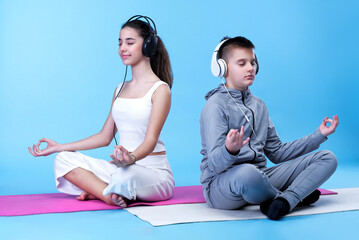 Two teenagers, friends from school, practicing yoga and meditation on their yoga mats, listening to relaxing music on their big headphones, blue background