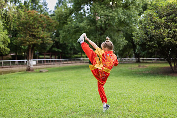 Cute little caucasian girl seven years old in red sport wushu uniform exercising in park at summer day. Lifestyle portrait of kung fu fighter child athlete