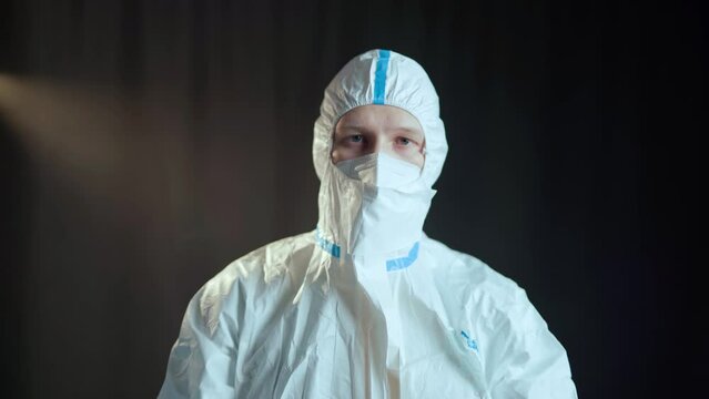 A man in a white medical suit of chemical protection and a white respirator and transparent medical gloves shows a gesture with his hands in the form of a cross, and then delivers boxing punche