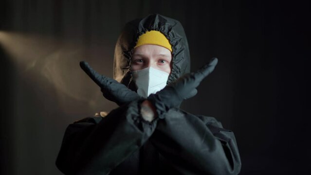 A man in a black medical chemical protection suit and a white respirator, wearing a yellow hat and black medical gloves shows a gesture with his hands in the form of a cross
