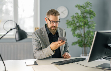 Caucasian businessman in stylish suit and eyewear using smartphone while sitting at office. Computer and tablet on table. Modern gadgets for work.