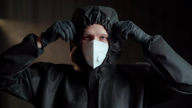 A man in a black medical chemical protection suit and a white respirator and black medical gloves removes the hood, and then the respirator and inhales deeply