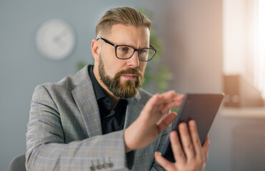 Successful businessman in stylish suit and eyeglasses sitting at modern office and looking on tablet screen with serious facial expression. Working process of busy person.