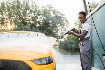 Handsome African young man in protective overalls, washing his yellow car manually with water high-pressure hose at outdoor self wash service. Car washing concept.