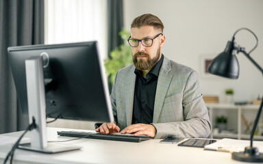 Fototapeta na wymiar Confident bearded man sitting at office desk and using modern pc for work. Handsome male person wearing stylish business suit and eyeglasses.
