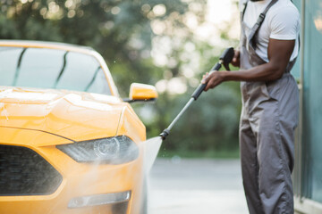 Close up cropped horizontal image of hands of African man washing his yellow luxury car using high pressure water, at outdoors car self washing service.