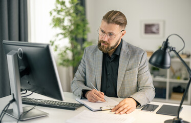 Bearded caucasian man in suit and eyeglasses taking notes while sitting at desk and looking on computer screen. Mature businessman with full concentration working at office.