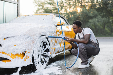 Car wash outdoors concept. Luxury modern car in foam. Young African man with high pressure jet...