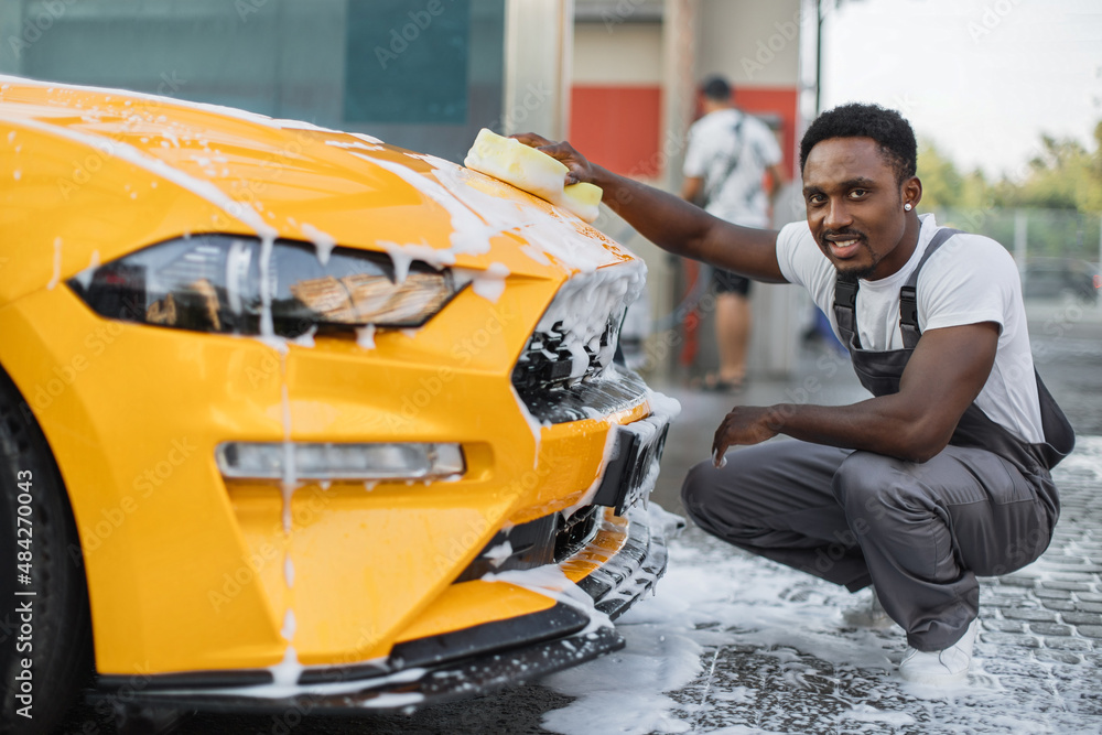 Poster Luxury modern sport car in soap foam outdoors at car wash service. Side view of handsome young African man using yellow sponge for cleaning rims outdoors.