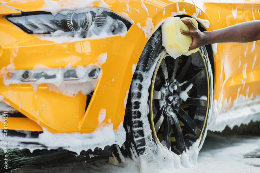 Wall mural Hands of African man holding yellow sponge, washing car wheel with foam. Cleaning of modern rims of luxury yellow car at self car wash service outdoors.